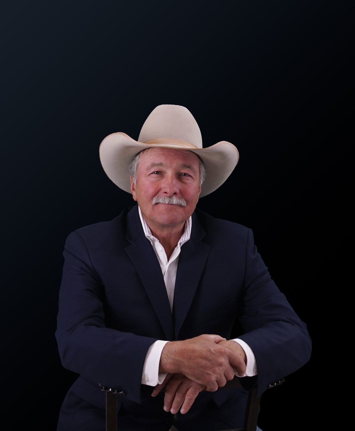D. Michael Thomas seated in a pale cowboy hat and dark jacket, hands folded over the back of a chair