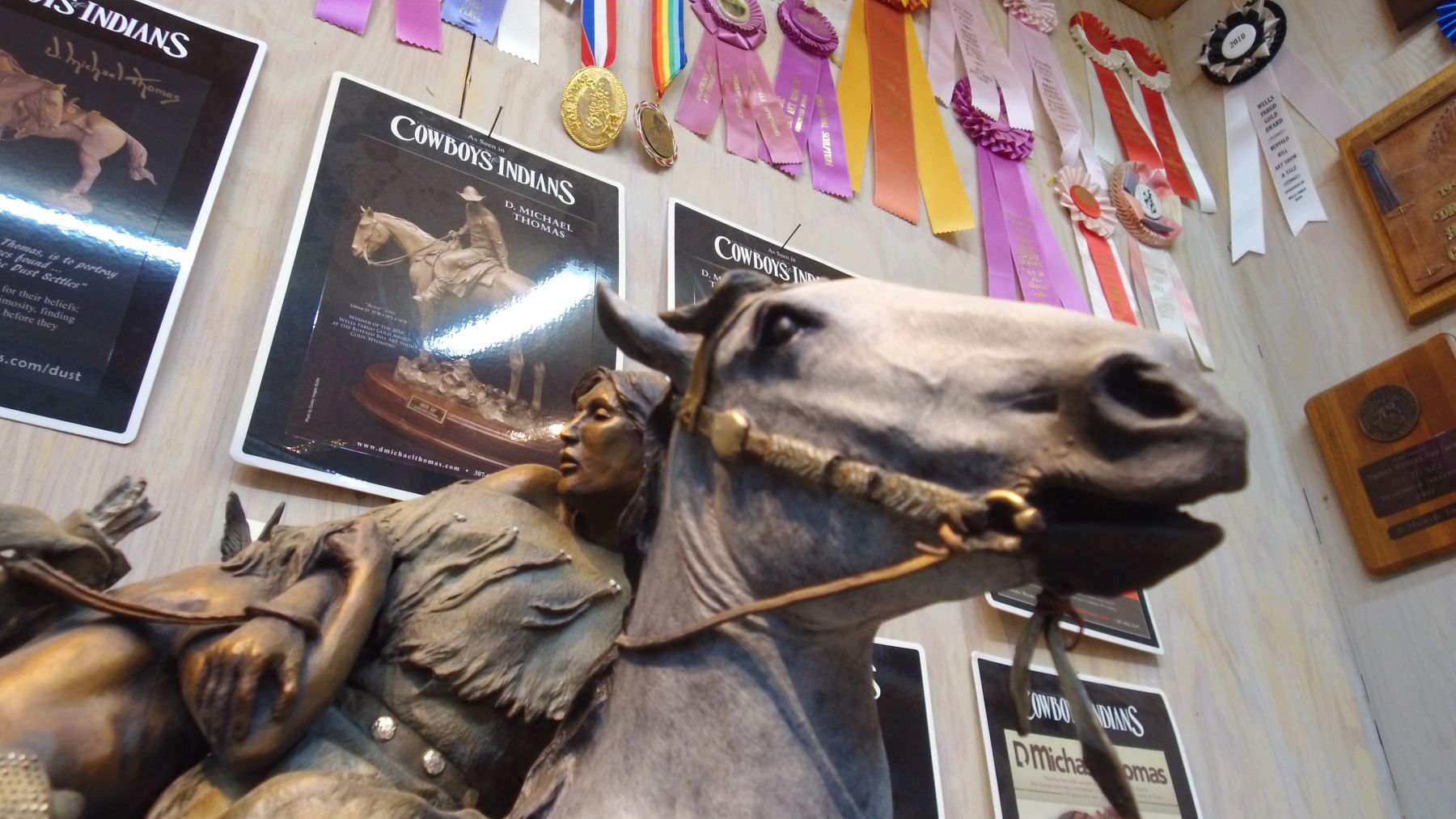 A mounted rider in bronze in the studio, below a wall of show ribbons and press